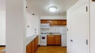 This single-floor unit offers a practical and well-organized layout designed for comfortable everyday living. The kitchen is a standout feature, equipped with warm honey-toned wood cabinetry, granite-style countertops, a full suite of white appliances including a refrigerator, electric range, over-the-range microwave, and a single-basin sink. An additional countertop peninsula provides extra prep space and a natural transition into the adjacent living area. Throughout the unit, light hardwood flooring creates a cohesive and warm aesthetic. The main living area is generously sized, featuring a textured ceiling and a large sliding glass door that invites abundant natural light and provides access to an outdoor space. A secondary room features a window with a curtain, painted cinder block walls, and the same hardwood flooring, lending it a clean, versatile character. The bathroom includes a full soaking tub with a shower, a curtain rod, grab bar safety features, and a warm wood vanity. The overall ambiance of the unit is clean, neutral, and functional, with consistent finishes throughout that create a unified and practical living environment.
