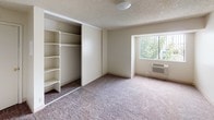 This single-floor space offers a straightforward and functional layout designed to maximize practicality. The main room is finished with wall-to-wall carpeting in a neutral tone, creating a soft and comfortable underfoot feel throughout. A prominent built-in closet spans a significant portion of one wall, featuring sliding panel doors and an interior organizer system complete with multiple shelving units and double hanging rods — providing ample storage and organization options. The walls are painted in a light, neutral beige tone that contributes to a clean and open atmosphere. The textured popcorn ceiling adds a classic finishing detail overhead. A wall-mounted HVAC unit is conveniently positioned beneath the window, ensuring climate control directly within the room. The window itself is fitted with a security bar grille and allows natural light to filter into the space while framing a view of lush outdoor greenery beyond. The overall ambiance of the space is calm and understated, with a focus on utility and livability. The neutral palette throughout makes it a versatile canvas well-suited for a variety of furnishing styles and personal touches.