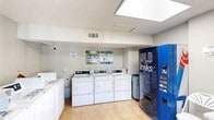 Located at 964 Simpson Terrace in Bedford, TX, this shared laundry facility offers a practical and functional space designed to serve residential users efficiently. The single-floor layout is finished with light-toned ceramic tile flooring and neutral gray walls, creating a clean, utilitarian environment that is easy to maintain.

The main area houses a row of front-load dryers paired with top-load washers along one wall, providing ample capacity for multiple users. Washer hookups are clearly visible along the adjacent wall, indicating additional machine capacity. A large cold drinks vending machine is conveniently positioned near the center of the room, offering refreshments during laundry sessions. A utility trash bin sits nearby for easy waste disposal.

A folding table with seating is available, giving users a surface to sort and fold laundry comfortably. The entryway features a windowed door that allows natural light to filter into the space, complemented by overhead fluorescent lighting and a skylight panel that brightens the interior. A textured ceiling adds a classic finish to the room.

Safety is addressed through a mounted fire extinguisher and an active security camera, with posted signage reminding users they are on camera. Instructional guides for washer use are posted on the wall, and an HVAC vent ensures adequate climate control. The space is accessed through a single exterior door, making it straightforward and accessible for all residents.