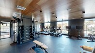 This contemporary fitness center is designed to deliver a premium workout experience within a thoughtfully crafted environment. The space is defined by its standout warm-toned wood panel ceiling, which contrasts beautifully against the dark charcoal walls and durable rubber flooring, creating an atmosphere that is both sleek and inviting.

The cardio zone lines the windowed perimeter of the space, where a row of treadmills, elliptical machines, and stationary bikes — including TRUE Fitness branded equipment — are arranged to take full advantage of the large windows. These floor-to-ceiling windows flood the space with natural light and offer open views of the surrounding treetops and cityscape beyond.

The strength training area is equally well-appointed, featuring a multi-functional cable and weight machine, a full rack of dumbbells ranging across multiple weights, barbells with plate storage, and an adjustable weight bench. Full-length mirrors line portions of the walls, enhancing the sense of openness and allowing users to monitor their form throughout their workouts.

Glass-paned interior doors connect seamlessly to adjacent areas, maintaining visual flow throughout. The combination of abundant natural light, high-end equipment, refined finishes, and a spacious open floor plan makes this fitness center a standout amenity space.