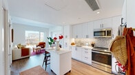 This thoughtfully designed one-bedroom, one-bathroom residence offers a smart and efficient open-concept floor plan with contemporary finishes throughout. The kitchen serves as a central hub of the space, outfitted with crisp white cabinetry extending to the ceiling, stainless steel appliances including a range, microwave, and refrigerator, and a mosaic tile backsplash that adds texture. A white quartz-topped island with bar seating provides additional prep and dining space, flowing seamlessly into the living area. The living room is bright and airy, anchored by large floor-to-ceiling windows that frame urban cityscape views and flood the room with natural light. Light-toned hardwood flooring runs throughout, complementing the clean white walls. The bedroom is generously proportioned, featuring an oversized window with sweeping city views, creating a bright and serene retreat. The bathroom includes a soaking tub with a clean white tile surround accented by a mosaic tile column and matte black fixtures, offering a sleek, modern aesthetic. A dedicated closet provides practical storage. The overall ambiance is modern and light-filled, with an efficient layout that makes excellent use of every square foot.