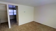 This two-floor space presents a clean, functional layout with neutral cream-colored walls that create a bright and open atmosphere. The main living area is finished with wall-to-wall carpeting in a warm tan tone, providing a comfortable underfoot feel. A partial wall divides the space, adding subtle architectural definition between areas while maintaining an open feel. A wall-mounted thermostat is conveniently positioned for climate control access. Adjacent to the main living area, a connected space features tile flooring and is illuminated by natural light streaming through a window, offering a practical contrast in flooring materials. A floor-level air vent is integrated into the base of the partial wall, ensuring efficient airflow throughout. The overall palette is understated and versatile, with white ceilings and neutral finishes that offer a blank canvas adaptable to a variety of interior styles. The transition between the carpeted and tiled areas suggests distinct functional zones within the home, contributing to a well-organized flow across the main level.