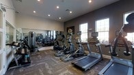 This fitness center is a thoughtfully designed workout facility built to support a comprehensive exercise routine. The space is outfitted with a row of modern cardio machines, including treadmills and elliptical trainers, all positioned to maximize usability and flow throughout the room. Large windows along one wall flood the space with natural light, creating an open and energizing atmosphere ideal for working out. A full-length mirror wall runs along the opposite side, visually expanding the room while allowing users to monitor their form during exercise. The ceiling features recessed lighting that supplements the natural light and ensures the space remains well-lit at all hours. The walls are finished in a deep, neutral tone that gives the room a sleek, modern aesthetic, complemented by durable, patterned flooring designed to withstand high-traffic use. Motivational artwork is displayed on the accent wall, adding a purposeful character to the environment. The overall layout is clean and organized, with ample space between equipment to allow for comfortable movement. This fitness center combines functional design with a polished, professional ambiance suited for daily training.