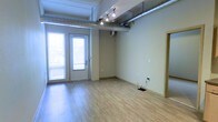 This single-floor space showcases a clean, open layout with a distinctive blend of industrial and contemporary design elements. Exposed metallic ductwork runs prominently along the ceiling, lending the space an urban, loft-inspired character. Adjustable track lighting mounted on the ceiling provides versatile illumination, complementing the natural light that streams in through a glass-paneled double door along one wall.

The flooring consists of light-toned hardwood-style planks that enhance the brightness of the interior and add warmth to the overall aesthetic. The walls are finished in a neutral, light beige tone, contributing to an open and airy atmosphere. Electrical outlets and data ports are positioned along the walls, suggesting practical utility for a variety of uses.

A secondary door on the adjacent wall provides additional access or connectivity to neighboring spaces. The ceiling height, while not dramatically elevated, accommodates the industrial ductwork without feeling cramped. Overall, the space presents a versatile, well-lit environment suited for office, studio, retail, or creative use, offering a functional yet characterful setting.