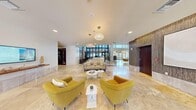 Located at 3498 E Ellsworth Ave in Denver's vibrant Cherry Creek neighborhood, this grand lobby hallway offers a sophisticated and welcoming first impression. The space is defined by its sweeping travertine tile flooring accented with rich hardwood inlay strips that guide visitors through the expansive interior. Soaring ceilings — dramatically double-height in key areas — amplify the sense of openness and grandeur throughout.

Natural light pours in through floor-to-ceiling glass panels and a prominent revolving door entry, connecting the interior to the surrounding streetscape. Two distinct chandelier styles punctuate the space: a cascading crystal bubble chandelier suspended from the double-height rotunda ceiling, and a pair of globe-style crystal pendant fixtures that illuminate a secondary seating zone.

Textured wood-panel accent walls add warmth and depth, contrasting elegantly with the smooth, neutral wall finishes. A curved architectural soffit with a dark ceiling detail defines the rotunda area, lending it a distinct identity within the larger floor plan. A reception desk with glass partition detailing anchors one end of the lobby, while multiple seating arrangements — featuring mustard-yellow armchairs, a curved gray sofa, and contemporary white accent tables — create inviting pockets for gathering. The overall ambiance is modern, polished, and professional.