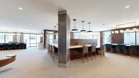 This generously scaled single-floor space masterfully blends modern design with warm, organic materials across a sweeping layout. The open common areas showcase rich chevron-patterned wood accent walls, tiled structural columns, and plush neutral carpeting that creates a cohesive, upscale atmosphere throughout. A striking central bar and kitchen area serves as the heart of the space, anchored by a large white quartz island, warm walnut cabinetry, decorative tile backsplash, and a cluster of pendant lights overhead. Exposed wood ceiling beams in the dining and living zones add rustic character, while floor-to-ceiling windows flood these areas with natural light and offer expansive outdoor views. A dedicated lounge features a dramatic floor-to-ceiling upholstered banquette with ambient underlighting, chevron-patterned dark walls, and classic black-and-white hexagonal tile flooring. A game room with patterned cement tile, a poker table, and additional recreational furnishings provides a playful retreat. Private workspaces include individual offices—one featuring a striking mural of the Alamo—offering focused environments for productivity. The bedroom provides an intimate, well-proportioned retreat, while the bathroom completes the well-rounded layout. Throughout, the design achieves a sophisticated balance of comfort, functionality, and refined aesthetic detail.