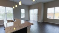 This single-floor residence showcases a seamlessly connected kitchen and living area designed for both functionality and comfort. The kitchen features a prominent granite-topped island with an integrated sink, offering ample prep and entertaining space. Pendant lights hang above the island, providing focused task lighting while adding a stylish focal point to the space.

The open-concept layout flows naturally into the living area, unified by rich, dark hardwood flooring throughout. Neutral gray walls create a versatile backdrop that complements the space's modern finishes. Natural light is a defining characteristic of this space, with a side window and a full-height sliding glass door dressed with vertical blinds drawing in abundant daylight and offering outdoor access.

A ceiling fan is positioned in the living area, contributing to year-round comfort. Clean baseboards and simple architectural lines give the space a contemporary feel, while the open sightlines between the kitchen and living areas create a sense of spaciousness. Overall, the unit delivers a practical, modern layout with cohesive design elements throughout.