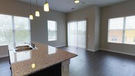 This single-floor residence showcases a seamlessly connected kitchen and living area designed for both functionality and comfort. The kitchen features a prominent granite-topped island with an integrated sink, offering ample prep and entertaining space. Pendant lights hang above the island, providing focused task lighting while adding a stylish focal point to the space.

The open-concept layout flows naturally into the living area, unified by rich, dark hardwood flooring throughout. Neutral gray walls create a versatile backdrop that complements the space's modern finishes. Natural light is a defining characteristic of this space, with a side window and a full-height sliding glass door dressed with vertical blinds drawing in abundant daylight and offering outdoor access.

A ceiling fan is positioned in the living area, contributing to year-round comfort. Clean baseboards and simple architectural lines give the space a contemporary feel, while the open sightlines between the kitchen and living areas create a sense of spaciousness. Overall, the unit delivers a practical, modern layout with cohesive design elements throughout.