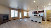 This two-floor home is designed with both comfort and functionality in mind, offering a thoughtfully planned layout that suits everyday living. The main level centers around an open-concept living and kitchen area that creates a seamless flow between spaces. The kitchen is well-equipped with dark-stained cabinetry, stainless steel appliances including a range and refrigerator, and a light-toned granite-style countertop island that doubles as a casual dining or prep surface. Pendant lighting hangs above the island, while recessed ceiling lights and a chandelier-style fixture over the dining area provide warm, layered illumination throughout the space. The living area features warm beige walls, neutral carpet flooring that transitions to hardwood-style flooring near the kitchen, and large windows that invite an abundance of natural light. A glass-panel exterior door provides additional brightness and access to the outdoors. The smooth, flat ceiling enhances the clean, modern feel of the interior. The second floor extends the home's living capacity, offering additional private space. Overall, the home presents a welcoming, move-in-ready atmosphere with contemporary finishes and a practical open floor plan.