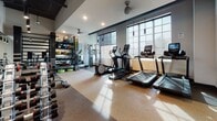 Located at 530 Meeting Street in Charleston, SC, this well-appointed fitness center offers a comprehensive workout environment designed to support a full range of exercise routines. The space features soaring ceilings with exposed black ductwork, lending an industrial-modern aesthetic that is both functional and stylish. Pendant lighting and recessed fixtures provide ample illumination throughout, complemented by an abundance of natural light streaming in through large, multi-pane windows along the exterior walls. The cardio zone is equipped with multiple treadmills and elliptical machines, each featuring integrated digital displays. A dedicated free weights area includes a full rack of dumbbells of varying sizes, accompanied by adjustable benches and a large wall mirror that visually expands the space and aids in form monitoring. Additional functional training equipment, including medicine balls, resistance tools, and a multi-station rack, is organized along one wall for easy access. The durable rubberized flooring throughout provides safety and comfort underfoot. Ceiling fans help maintain air circulation during intense workouts. A separate entry door offers convenient, controlled access. The overall layout is open and thoughtfully organized, ensuring users can move freely between equipment zones.