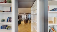 This elegantly designed single-floor space is a masterclass in contemporary interior styling, combining comfort and refined aesthetics throughout its interconnected zones. The herringbone-patterned hardwood flooring serves as a unifying element across the entire layout, lending warmth and visual continuity to every area.

At the heart of the space is a striking full-service bar, anchored by a long granite-topped counter with built-in fixtures and lined with upholstered bar stools. Above, a sculptural chandelier with cascading organic elements adds a dramatic focal point, while recessed lighting complements the ambient glow. A textured feature wall and curated artwork enhance the bar's sophisticated character.

The lounge area accommodates generous sectional sofas arranged around a bold multi-panel world map artwork set against a metallic-finish wall, accented by exposed architectural beams that add depth to the ceiling. An adjoining sitting room offers a more intimate setting with curved club chairs, brass accent tables, and a weathered wood-panel feature wall adorned with an eye-catching portrait. Built-in bookshelves line multiple walls throughout, providing both functional storage and visual interest. A transitional hallway connects the zones, featuring the same distinctive wall textures and thoughtful architectural detailing that define this cohesive, upscale space.