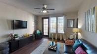 This single-floor unit offers a thoughtfully designed layout that balances comfort and functionality across its key living spaces. The living room serves as the heart of the home, featuring rich hardwood-style flooring, a ceiling fan, and a wall-mounted television. A set of French doors flanked by large windows floods the space with natural light and provides access to an outdoor area, while neutral-toned walls create a versatile backdrop for the room's furnishings.

The kitchen is outfitted with dark espresso cabinetry, light countertops, and a full suite of black appliances including an electric range, built-in microwave, dishwasher, and a full-size refrigerator. A hallway off the kitchen leads to an in-unit stacked washer and dryer, adding everyday convenience. The kitchen flows openly toward the living area, supported by a breakfast bar that enhances the sense of connectivity throughout the space.

The bedroom is generously sized, benefiting from multiple windows that bring in ample natural light. A ceiling fan adds comfort, and the room features plush carpeting for a cozy feel. The bathroom includes a full soaking tub and shower combination with earth-toned tile surround and dark cabinetry, offering a clean and functional design. Overall, the unit presents a cohesive, modern aesthetic throughout.