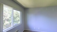 This single-story space features a simply designed room finished with soft grey-blue painted walls that create a calm, neutral atmosphere. The room is carpeted, adding warmth underfoot, and is framed by clean white baseboards that provide a crisp contrast to the walls. A large sliding window on one wall allows ample natural light to fill the space while offering pleasant views of mature, leafy trees outside, bringing a sense of connection to the outdoors. The textured popcorn ceiling adds a classic touch characteristic of traditional residential construction. A wall-mounted heating unit sits at the base of the exterior wall beneath the window, providing climate control directly within the room. Electrical outlets are conveniently positioned along the walls. The room is currently unfurnished, making it easy to appreciate its clean lines and versatile layout. The overall ambiance is quiet and understated, well-suited for use as a bedroom, home office, or flex space. The abundance of natural light and the greenery visible through the window contribute to a pleasant and inviting environment.