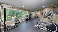Located at 601 Jones Ferry Rd in Carrboro, NC, this dedicated fitness center offers a well-appointed exercise room designed to support a comprehensive workout experience. The space features durable rubber flooring throughout, providing both safety and comfort underfoot. Recessed lighting illuminates the room evenly, complemented by an abundance of natural light streaming in through large windows and a sliding glass door that overlooks lush, tree-lined surroundings — creating an inviting and energizing atmosphere.

The room is equipped with a diverse array of fitness equipment, including a treadmill, elliptical trainer, stationary recumbent bike, and a rowing machine for cardio training. Strength training is well supported by a multi-station cable machine, an adjustable weight bench, and a dumbbell rack with a full set of free weights. A stability ball and additional accessories round out the offerings. A wall of mirrors on one side enhances the sense of space and allows users to monitor their form during workouts.

The neutral, warm-toned walls are accented by a soft blue horizontal stripe, adding a subtle decorative touch. A mounted flat-screen television and a safety station on the wall further contribute to the room's functionality. The glass door provides direct outdoor access, allowing for easy ventilation and a seamless connection to the exterior.