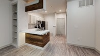 This well-appointed single-floor unit offers a clean, contemporary design highlighted by light wood-style vinyl flooring and a consistent neutral color palette that flows seamlessly throughout the space. The open-concept kitchen serves as a central feature, showcasing gray shaker-style cabinetry, white quartz countertops, a subway tile backsplash, and a full suite of stainless steel appliances including a range, microwave, dishwasher, and refrigerator. A peninsula island with a warm wood accent finish and built-in sink adds both style and functionality, while recessed lighting illuminates the space efficiently. The kitchen connects directly to the entryway, which features a paneled front door and an open sightline into the living areas. The living and dining areas are bright and spacious, benefiting from multiple windows dressed with horizontal blinds that allow for ample natural light. Crown molding and a ceiling fan add refined detail to the living area. The bedroom is generously sized with tall walls, dual windows, and the same consistent flooring. The bathroom is tastefully finished with a gray vanity cabinet, white countertop, large mirror, and wood-style flooring, maintaining the cohesive design aesthetic found throughout the unit.