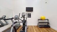 The Tides Fitness Center at 13517 W Glendale Ave in Glendale, AZ offers a well-equipped, single-level workout facility designed to support a variety of exercise routines. The space features warm wood-look flooring, crisp white walls, and notably high ceilings that lend an open, airy atmosphere. A large wall-to-wall mirror along one side of the room enhances the sense of space and allows users to monitor their form during workouts. Natural light filters in through a row of elevated clerestory windows, complementing the bright interior.

The cardio section is outfitted with Matrix-brand treadmills, an elliptical trainer, and a recumbent exercise bike, catering to users of varying fitness levels. A wall-mounted flat-screen television provides entertainment during workouts. For strength training, the facility includes a multi-function cable and weight machine, offering a range of resistance exercises. A well-organized equipment rack stores an assortment of stability balls, resistance bands, foam rollers, and free weights, supporting functional fitness and stretching routines. Double doors provide easy entry and exit, and posted fitness center rules ensure a safe and orderly environment. The overall design is clean, functional, and conducive to an effective workout experience.