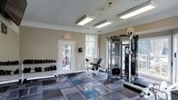 This fitness center offers a practical and well-organized workout environment designed to accommodate a variety of exercise routines. The space is finished with neutral warm-toned walls, crisp white crown molding, and a durable patterned tile floor in shades of blue and gray, providing both functionality and visual appeal.

Natural light fills the room through multiple white-framed windows, complemented by overhead fluorescent panel lighting that ensures consistent brightness throughout. A glass-paneled exterior door offers additional light and convenient access to the outdoors.

The equipment layout is thoughtfully arranged to maximize the available floor space. A tiered dumbbell rack lines one wall, stocked with a full range of free weights for strength training. At the center of the room, a weight bench and leg press machine provide targeted resistance training options. A tall, multi-functional cable machine anchors one side of the space, supporting a wide range of cable-based exercises.

A security camera and fire safety exit signage reflect attention to safety standards throughout the facility. The overall ambiance is clean and functional, making this fitness center a capable and inviting space for residents or members looking to maintain an active lifestyle.