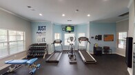 Located at 1083–1085 NE Green Oaks Blvd in Arlington, Texas, this fitness center is a single-floor facility thoughtfully organized into distinct workout zones, offering a comprehensive range of exercise options. The space features light blue and grey walls accented with motivational wall decals, including a bold "Just Don't Quit" graphic, creating an energetic and encouraging atmosphere throughout.

The cardio zone is equipped with multiple treadmills and an elliptical machine, complemented by a wall-mounted television for entertainment during workouts. Natural light filters in through several windows fitted with horizontal blinds, contributing to a bright and open feel. Recessed lighting and crown molding add a polished, finished quality to the interior.

A dedicated free weights area includes a tiered dumbbell rack with a full range of weights, an adjustable workout bench, and additional resistance accessories. The strength training section houses a multi-functional cable machine suitable for a wide variety of exercises, alongside decorative fitness-themed wall graphics.

A separate spin and cycling zone accommodates multiple stationary bikes and an upright cycle, with large windows providing ample natural light and outdoor views. A battle rope is also available for functional training. Rubber flooring throughout the facility ensures safety and durability, making this a well-rounded and inviting fitness destination.