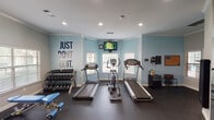 Located at 1083–1085 NE Green Oaks Blvd in Arlington, Texas, this fitness center is a single-floor facility thoughtfully organized into distinct workout zones, offering a comprehensive range of exercise options. The space features light blue and grey walls accented with motivational wall decals, including a bold "Just Don't Quit" graphic, creating an energetic and encouraging atmosphere throughout.

The cardio zone is equipped with multiple treadmills and an elliptical machine, complemented by a wall-mounted television for entertainment during workouts. Natural light filters in through several windows fitted with horizontal blinds, contributing to a bright and open feel. Recessed lighting and crown molding add a polished, finished quality to the interior.

A dedicated free weights area includes a tiered dumbbell rack with a full range of weights, an adjustable workout bench, and additional resistance accessories. The strength training section houses a multi-functional cable machine suitable for a wide variety of exercises, alongside decorative fitness-themed wall graphics.

A separate spin and cycling zone accommodates multiple stationary bikes and an upright cycle, with large windows providing ample natural light and outdoor views. A battle rope is also available for functional training. Rubber flooring throughout the facility ensures safety and durability, making this a well-rounded and inviting fitness destination.