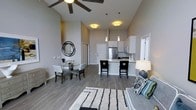 This single-floor residence showcases a thoughtfully designed open-concept layout that seamlessly integrates the living, dining, and kitchen areas into one cohesive and airy space. The most striking architectural feature is the soaring vaulted ceiling, which creates an impressive sense of volume and grandeur throughout the main living area. A ceiling fan with sleek, broad blades is centrally positioned to complement the height of the room.

The kitchen is both functional and stylish, equipped with stainless steel appliances, white cabinetry, and a peninsula island with bar seating — ideal for casual dining or entertaining. Pendant lighting hangs above the island, adding warmth and a contemporary touch. The dining area is situated adjacent to the kitchen and features a round glass-top table with upholstered chairs, accented by a decorative circular wall mirror.

The living area is open and inviting, with a neutral color palette of warm grays and whites that lends a clean, modern aesthetic throughout. Wide-plank hardwood-style flooring runs across the entire main floor, tying the spaces together with a consistent, sophisticated finish. Natural light filters in through nearby windows, enhancing the bright and welcoming ambiance of this well-appointed space.
