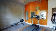 This fitness center combines industrial-inspired design with vibrant color to create an energizing workout environment. The space is defined by a striking contrast between raw, exposed concrete walls and bold orange accent walls, giving the room a dynamic and motivating atmosphere. Visible overhead piping adds to the industrial character of the space, while the angled ceiling lends architectural interest.

The room is equipped with key cardio machines, including a treadmill and a Concept2 rowing machine, positioned to make efficient use of the available floor space. Rubberized flooring provides a durable, safe surface for exercise. Mirrors are strategically mounted on the orange walls, serving both functional and aesthetic purposes by enhancing the sense of space and allowing users to monitor their form during workouts. A decorative clock anchors the main accent wall, adding a practical yet stylish touch. Natural light filters in through a window, contributing to an open and welcoming feel. Overall, the fitness center offers a well-appointed, purposefully designed space for residents to maintain an active lifestyle.