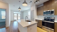 This single-floor space showcases a contemporary open-concept design that seamlessly connects the kitchen and living areas. The kitchen is a standout feature, centered around a white quartz island with an undermount sink and stylish industrial pendant lighting overhead. Rich wood-tone cabinetry extends from base to upper cabinets, complemented by a classic subway tile backsplash and a full suite of stainless steel appliances, including a refrigerator, range, and microwave. Track lighting above the kitchen provides additional task illumination.

Warm hardwood-style flooring flows throughout the main living and kitchen areas, creating a cohesive and inviting atmosphere. Large windows along the exterior walls allow natural light to fill the open living space, which also features a ceiling fan for added comfort. The layout, as seen in the floor plan view, is efficiently organized with clear sightlines from the living area through the kitchen.

The bedroom offers a quieter retreat with neutral carpet flooring and clean white walls. The bathroom is finished with wood-look flooring, a soaking tub with white tile surround, a granite-topped vanity with wood cabinetry, and direct access to a walk-in closet with built-in shelving, adding practical storage to the space.