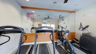 This fitness center is thoughtfully designed to provide an energizing and functional workout environment. The space features bold orange accent walls paired with crisp white trim, creating a vibrant atmosphere that complements the motivational wall decals — including "Just Don't Quit" and "Commit 2 B Fit" — displayed prominently across the large floor-to-ceiling mirrored wall. The mirrors not only reinforce the motivational theme but also visually expand the room, giving it an open and spacious feel. A ceiling fan ensures comfortable air circulation during workouts. The facility is equipped with a range of cardio machines, including treadmills, a recumbent bike, and an elliptical-style trainer, offering users a variety of workout options. Natural light filters in through a window, brightening the space and contributing to an uplifting exercise environment. Recessed lighting supplements the natural light for consistent illumination throughout. The dark rubberized flooring provides a durable, slip-resistant surface well-suited for fitness activities. Overall, the space is clean, purposeful, and designed to motivate users at every fitness level.
