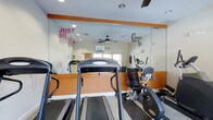 This fitness center is thoughtfully designed to provide an energizing and functional workout environment. The space features bold orange accent walls paired with crisp white trim, creating a vibrant atmosphere that complements the motivational wall decals — including "Just Don't Quit" and "Commit 2 B Fit" — displayed prominently across the large floor-to-ceiling mirrored wall. The mirrors not only reinforce the motivational theme but also visually expand the room, giving it an open and spacious feel. A ceiling fan ensures comfortable air circulation during workouts. The facility is equipped with a range of cardio machines, including treadmills, a recumbent bike, and an elliptical-style trainer, offering users a variety of workout options. Natural light filters in through a window, brightening the space and contributing to an uplifting exercise environment. Recessed lighting supplements the natural light for consistent illumination throughout. The dark rubberized flooring provides a durable, slip-resistant surface well-suited for fitness activities. Overall, the space is clean, purposeful, and designed to motivate users at every fitness level.