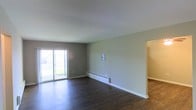 This single-floor space presents a clean, straightforward layout centered around a spacious main living area. Dark hardwood flooring runs throughout, offering a warm contrast against the neutral gray-toned walls and bright white trim. A sliding glass door along one wall serves as the room's primary source of natural light, framing a view of a grassy outdoor area and lending the interior an open, airy quality.

A baseboard heating unit runs along the lower portion of the far wall, providing a practical, unobtrusive climate control solution. A wall-mounted panel, likely a thermostat or electrical component, is positioned nearby. White baseboard trim lines the perimeter of the room, adding a crisp finishing detail to the otherwise minimal aesthetic.

An open doorway on the right side of the main room leads to an adjacent hallway or secondary space, suggesting a connected layout with additional rooms beyond the main living area. The overall ambiance of the space is neutral and understated, with a move-in-ready quality that allows for flexible furnishing and personalization. The combination of natural light, hard flooring, and simple finishes gives the space a practical yet inviting character.