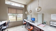 Located at 1550 Crescent Pointe Pkwy in College Station, Texas, this single-room business center office offers a thoughtfully designed workspace that maximizes functionality within a compact footprint. The room is accessed through a glass-paneled door framed by a carpeted entry corridor, with a wall-mounted directory panel and built-in display case lending a polished first impression.

Inside, the office features multiple built-in workstations arranged along opposing walls, each equipped with desktop computers, keyboards, and privacy screens to support focused individual work. Rich dark-wood cabinetry with light countertops provides ample storage and surface space, while ergonomic task chairs in both classic black and warm orange add a contemporary touch.

The room benefits from soaring ceiling height, which creates an open, airy feel despite its compact size. A row of globe-style pendant lights hangs at varying heights, casting warm, even illumination throughout. Natural light filters in through a large clerestory window overlooking a landscaped exterior, enhancing the bright and welcoming atmosphere.

Neutral beige walls serve as a clean backdrop for a striking series of colorful, multi-panel abstract artworks, injecting energy and creativity into the professional environment. Patterned carpeting in muted tones grounds the space and ties together its modern, refined aesthetic.
