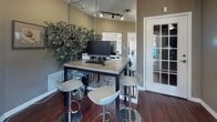 This single-floor community clubhouse offers a thoughtfully designed space that balances functionality with an inviting atmosphere. Rich hardwood flooring flows throughout, anchoring the warm, taupe-toned walls that lend a cohesive and polished look to every area.

The reception area serves as the heart of the space, featuring a dark wood information desk stocked with reading materials, a ceiling fan overhead, and large windows that draw in generous natural light and views of the surrounding greenery. Track lighting adds focused illumination throughout, complementing the natural brightness.

French-paned glass partition walls connect the main areas while maintaining a sense of openness, allowing sightlines into the adjacent lounge. The lounge area is furnished with comfortable seating, a vibrant teal accent wall, and additional natural light streaming through glass doors.

The business center is a standout feature, equipped with computer workstations set atop a high communal table surrounded by modern chrome bar stools. A full-length mirror on the wall amplifies the sense of space, while framed artwork and decorative plants add a refined, welcoming touch. Wi-Fi access is provided, making this an ideal spot for productivity. Together, these areas create a versatile, community-oriented environment designed for both work and relaxation.