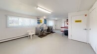 Located at 1950 Sheridan Dr in Buffalo, NY, this single-floor utility room serves as a shared laundry and storage facility. The space is cleanly finished with white walls, smooth concrete flooring, and fluorescent overhead lighting that ensures bright, even illumination throughout.

The laundry area occupies one side of the room and is equipped with multiple commercial-grade front-loading washing machines and dryers, accompanied by a utility sink and instructional signage posted on the walls. Vertical blinds on the windows allow natural light to filter into this section, adding to the functional atmosphere. A seating area with a chair provides a convenient waiting spot during laundry cycles.

On the opposite side of the room, a row of numbered individual storage units lines the walls, offering organized, dedicated storage spaces. The units feature white panel doors and extend along two walls, maximizing storage capacity within the room.

A bold red multi-tiered work or folding table stands prominently in the central area, serving as a practical surface for sorting and organizing. Additional amenities include a fire extinguisher mounted on the wall, an intercom or utility panel, and a trash receptacle. A "No Exit" door is also visible, emphasizing the room's layout as a controlled-access utility space. Overall, this well-organized facility provides essential shared services in a straightforward, practical setting.