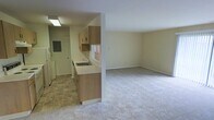 This single-floor residential unit offers a practical and open layout that connects the kitchen and living areas seamlessly. The kitchen is equipped with light wood cabinetry, a countertop with an integrated sink, and includes appliances such as a refrigerator and stove. Tile flooring in the kitchen provides a durable and easy-to-clean surface, while a window above the sink allows for natural light to filter into the space. An electrical panel is conveniently located near the kitchen entrance. A half-wall partition subtly defines the boundary between the kitchen and the living area, maintaining an open and airy feel throughout. The living area is generously sized, featuring wall-to-wall carpeting that adds warmth and comfort underfoot. Neutral-toned walls throughout the unit create a clean, versatile backdrop suitable for a variety of interior styles. Flush lighting fixtures provide consistent illumination across both areas. The overall ambiance is simple, functional, and move-in ready, making it a practical living space with room to personalize.
