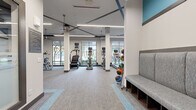 Located at 2350 8th Avenue South in Nashville, TN, this fitness center offers a thoughtfully designed, single-floor layout encompassing two distinct exercise rooms and a support area. The main exercise room is a spacious, open-concept cardio and strength training floor bathed in natural light from large floor-to-ceiling windows that look out onto the surrounding greenery. Soaring ceilings with bold geometric teal and dark accent details lend the space a dynamic, modern aesthetic, while linear pendant and recessed lighting ensures excellent visibility throughout. The cardio zone is well-stocked with stair climbers, ellipticals, and treadmills arranged along the windowed wall, while the strength area features a multi-station cable machine, a full dumbbell rack ranging across multiple weights, a WaterRower, and ample open floor space for functional training. Medicine ball storage and built-in bench seating with cubbies provide convenient organization near the entrance. A second, dedicated spin studio offers a more intimate setting, lined with floor-to-ceiling mirrors, ceiling fans, and multiple stationary cycling bikes. The consistent use of patterned carpet flooring with teal accent inlays ties the two rooms together, creating a cohesive and professional fitness environment.