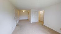 This single-floor residential space offers a straightforward and functional layout with a clean, neutral aesthetic throughout. The interior is finished with light beige wall-to-wall carpeting that runs seamlessly across the rooms, contributing to a soft and cohesive feel underfoot. The walls are painted in crisp white, lending an airy brightness to the space even in the absence of large windows in the main area.

A notable feature of the primary room is a built-in closet with double bifold doors, providing convenient and organized storage. Multiple doorways are visible throughout, connecting the main room to additional spaces — including what appears to be a hallway leading further into the unit and an exterior or secondary door, suggesting a practical circulation path through the home.

Overhead lighting fixtures are centrally placed, ensuring even illumination across the rooms. The overall layout appears open and unobstructed, making it well-suited for flexible furniture arrangements. The space presents a blank canvas with its minimal finishes and neutral palette, offering new occupants the opportunity to personalize and furnish according to their own style and needs.