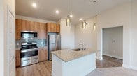 This single-floor unit offers a thoughtfully designed open-concept layout connecting the kitchen and living areas. The kitchen is a standout feature, equipped with warm-toned wood cabinetry extending to the ceiling, stainless steel appliances including a range, microwave, and refrigerator, and a light blue glass tile backsplash. A central island with a granite countertop and undermount sink serves as the kitchen's focal point, topped with three elegant glass pendant lights that add a contemporary touch. Wood-style flooring runs throughout the kitchen and dining area, transitioning to plush carpet in the living space and bedroom.

The living area is bright and open, with a ceiling fan overhead and access through French doors to a private balcony that overlooks expansive open green space and distant cityscape views. The bedroom is a quiet, well-proportioned room with soft carpeting and neutral walls.

The bathroom features warm wood cabinetry with granite countertops, a large vanity mirror, wood-style flooring, and a glass-enclosed walk-in shower with tile surround. Throughout the unit, neutral tones, recessed lighting, and clean finishes create a modern and cohesive aesthetic.