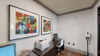 This business center at 8500 Franciscan Woods Dr in Columbus, Georgia, is a compact yet efficiently designed workspace that provides essential productivity amenities in a polished, professional setting. The room features warm hardwood-style flooring, crisp white walls accented by gray crown molding, and a clean, contemporary aesthetic throughout. A dark espresso-toned L-shaped desk serves as the focal point, equipped with a desktop computer, keyboard, and a laser printer, making it a fully functional workstation for everyday business needs. Bold, colorful framed artwork adorns the walls, adding a vibrant pop of personality to the otherwise neutral interior. The space is enclosed by floor-to-ceiling glass walls with sleek black metal framing, offering visibility into the surrounding amenity corridor while maintaining an open, airy feel. A hand sanitizer station is conveniently positioned within the room. Recessed lighting provides even, well-distributed illumination throughout. Views through the glass partitions reveal adjacent amenities, including a fitness center, enhancing the sense of connectivity within the broader facility. The space is thoughtfully appointed for focused work in a shared community environment.