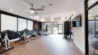 Located in Plano, TX, this fitness studio is a purpose-built exercise space designed to support a wide range of workout styles. The single-floor layout is finished with durable wood-look flooring throughout, providing a clean and functional foundation for high-energy activities. Dual ceiling fans maintain airflow across the room, complementing the recessed lighting that keeps the space well-lit at all times. Large floor-to-ceiling windows and a glass-paneled entry door flood the interior with natural light and offer views of the surrounding outdoor greenery, creating an open and energizing atmosphere. A bold dark accent wall serves as a focal backdrop, adorned with an illuminated neon "Make It Happen" motivational sign that sets the tone for the space. The studio is equipped with a row of stationary cycling bikes, a freestanding heavy punching bag and speed bag rig, a rack of medicine balls and stability balls, kettlebells, and a large wall-mounted flat-screen television. A mobile room divider offers flexible layout options. Glass partitions connect the studio to an adjacent fitness area stocked with additional cardio and strength equipment, a retail display, and a water fountain, making this a comprehensive fitness destination.