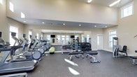 This fitness center is designed to support a comprehensive workout experience within a clean, modern facility. The space features soaring ceilings that lend an open, airy atmosphere, enhanced by recessed lighting and natural light streaming in through multiple windows along the walls. A full-length wall mirror spans the rear of the room, visually expanding the space and allowing exercisers to monitor their form during workouts. The cardio zone is well-appointed with a row of treadmills and elliptical machines, all positioned to face the mirrored wall and mounted flat-screen televisions for entertainment during exercise. The strength training area is stocked with a comprehensive dumbbell rack ranging across multiple weight increments, along with weight benches and various free weight accessories. A selection of medicine balls and stability equipment adds versatility for functional training. The flooring throughout is a durable, dark rubber surface ideal for high-impact activities and equipment use. The neutral color palette of warm whites and soft grays gives the facility a professional yet welcoming ambiance, making it suitable for users of all fitness levels.