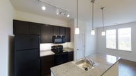 The Neely is a thoughtfully designed 1-bedroom, 1-bathroom residence that makes excellent use of its space through an open, functional floor plan. The kitchen serves as a central highlight, featuring dark espresso cabinetry that extends to the ceiling for ample storage, granite countertops, and black appliances including a refrigerator, electric range, and built-in microwave. A kitchen island with an integrated stainless steel sink and pendant lighting overhead adds both style and utility, creating a natural gathering point. Track lighting above the main kitchen area ensures the workspace is well-lit. The open sightlines connect the kitchen seamlessly to the living area, enhancing the sense of space and encouraging a flowing layout. A louvered closet door is visible just off the kitchen, hinting at practical storage solutions integrated throughout the unit. The overall aesthetic balances modern contrast — dark cabinetry against light countertops and neutral walls — delivering a clean, contemporary ambiance. The single-floor layout ensures easy accessibility, and the cohesive design choices throughout the space lend a polished, move-in-ready feel suited to comfortable everyday living.