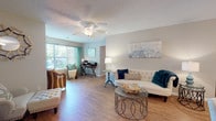 Located at 22 Colony Gardens Rd in Beaufort, South Carolina, this single-floor residence offers a thoughtfully designed layout with a warm and inviting coastal ambiance. The open-concept living area is anchored by wood-look flooring, a ceiling fan, and a textured ceiling, with generous windows dressed in teal curtains that flood the space with natural light and offer pleasant outdoor views. A sitting nook within the living area provides a comfortable retreat, complemented by a built-in desk area ideal for a home office setup.

The living space flows seamlessly into a dining area and kitchen, which features warm wood cabinetry, a peninsula breakfast bar with seating, a full suite of white appliances, and track lighting overhead. The open sightlines between these areas create a cohesive, functional common space well-suited for both everyday living and entertaining.

The residence includes multiple bedrooms, each finished with plush carpeting, ceiling fans, and windows that invite natural light. The primary bedroom offers a walk-in closet and ample space, while additional bedrooms provide versatility for guests or other uses.

The bathroom is neatly appointed with a wood-tone vanity, a tub-shower combination, and hardwood-style flooring, maintaining a consistent, clean aesthetic throughout the home.