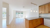 This well-designed single-floor residence offers a clean, contemporary aesthetic throughout, unified by light hardwood flooring and crisp white walls that create an airy, cohesive atmosphere. The open-concept kitchen anchors the living area with warm honey-toned cabinetry, granite countertops, a central island, and a full suite of white appliances including a gas range and side-by-side refrigerator. Pendant lighting and a track rail overhead illuminate the island, while the layout flows naturally into a generous living and dining area bathed in natural light. Sliding glass doors lead directly to a private balcony with an exterior railing, providing pleasant outdoor views of surrounding greenery. The bedrooms are well-proportioned, each featuring large windows that invite abundant sunlight and views of the tree-lined surroundings. One bedroom offers corner window placement, enhancing the sense of openness. A spacious walk-in closet with dual shelving provides excellent storage. The bathroom is finished with elegant marble-style tile flooring, a glass-enclosed walk-in shower with white subway tile, and clean, minimalist fixtures. The overall design emphasizes brightness, functionality, and modern comfort throughout every area of the space.