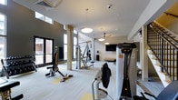 This two-floor fitness facility offers a thoughtfully designed workout environment that combines functionality with modern aesthetics. The space is bathed in natural light streaming through large floor-to-ceiling windows, creating an open and energizing atmosphere that enhances the exercise experience.

The gym is outfitted with professional-grade equipment, including Hoist strength-training machines, cable systems, and a variety of resistance and conditioning stations. The light-toned flooring provides a clean, neutral base that complements the contemporary two-tone wall design, featuring a combination of crisp white and deep charcoal grey accents.

Structural columns punctuate the space, adding an architectural element while defining workout zones. Overhead, modern pendant light fixtures and a ceiling fan contribute to a comfortable and well-lit environment. A wall-mounted flat-screen television is positioned for easy viewing during workouts. The high ceilings lend a sense of spaciousness and airiness throughout the facility.

Large glass entry doors provide direct access to the exterior, further connecting the interior to the outside environment. Security features, including overhead cameras, are also present. Overall, the space delivers a clean, professional, and welcoming fitness environment suited for a wide range of training needs.