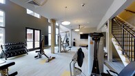 This two-floor fitness facility offers a thoughtfully designed workout environment that combines functionality with modern aesthetics. The space is bathed in natural light streaming through large floor-to-ceiling windows, creating an open and energizing atmosphere that enhances the exercise experience.

The gym is outfitted with professional-grade equipment, including Hoist strength-training machines, cable systems, and a variety of resistance and conditioning stations. The light-toned flooring provides a clean, neutral base that complements the contemporary two-tone wall design, featuring a combination of crisp white and deep charcoal grey accents.

Structural columns punctuate the space, adding an architectural element while defining workout zones. Overhead, modern pendant light fixtures and a ceiling fan contribute to a comfortable and well-lit environment. A wall-mounted flat-screen television is positioned for easy viewing during workouts. The high ceilings lend a sense of spaciousness and airiness throughout the facility.

Large glass entry doors provide direct access to the exterior, further connecting the interior to the outside environment. Security features, including overhead cameras, are also present. Overall, the space delivers a clean, professional, and welcoming fitness environment suited for a wide range of training needs.