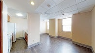 This single-floor space presents a clean, functional layout suited for a variety of uses. The main open area is finished with dark hardwood-style flooring that contrasts with the warm beige walls, creating a neutral and practical aesthetic. A drop ceiling with recessed lighting and a grid-tile design provides even illumination throughout the space, while a smoke detector and sprinkler system are also integrated overhead.

Natural light enters through two double-hung windows positioned along the far wall, brightening the interior and offering views to the outside. White baseboards line the perimeter of the room, adding a clean finishing detail to the walls.

To one side, a partial wall divides the main area from a kitchenette alcove, which features dark wood cabinetry, a countertop, and a sink, providing basic utility functions. The open floor plan allows for flexible arrangement and use of the space.

Overall, the unit offers a straightforward, well-maintained environment with practical finishes, functional amenities, and a layout that balances open living space with a dedicated utility area.
