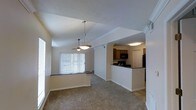 This single-floor residence showcases a thoughtfully designed open-concept layout that seamlessly connects the living, dining, and kitchen areas. The living and dining space is anchored by vaulted ceilings that lend an airy, expansive feel to the interior. Soft carpeted flooring runs throughout, adding warmth and comfort underfoot, while neutral-toned walls create a clean, welcoming ambiance. A ceiling fan and a stylish pendant light fixture hang from the vaulted ceiling, providing both function and a decorative touch. Large vertical blinds cover a sliding glass door or wide window, allowing for natural light to filter in while offering privacy. The kitchen is positioned adjacent to the main living area and is defined by a raised breakfast bar peninsula with dark countertops, warm wood-toned cabinetry, and black appliances including a microwave and refrigerator. Crown molding details add a refined architectural accent throughout. The open sightlines between spaces make the overall layout feel cohesive and well-connected, ideal for everyday living and entertaining alike.