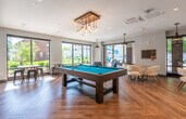 This contemporary single-floor amenity space is thoughtfully designed to offer a dynamic blend of recreation, relaxation, and productivity. The open layout is anchored by warm hardwood flooring throughout and filled with natural light streaming in through expansive floor-to-ceiling windows that frame views of a well-maintained outdoor landscape with manicured greenery.

At the heart of the space sits a full-size pool table beneath a striking rustic wood-and-Edison-bulb chandelier, creating a focal point for social interaction. Adjacent to it, multiple seating vignettes offer versatile lounging options — including modern white swivel chairs around casual tables and a cozy corner nook with teal upholstered armchairs positioned to enjoy the outdoor views.

A dedicated lounge area features a warm wood-paneled ceiling, a large wall-mounted flat-screen TV, a bold geometric-patterned area rug, and a mix of navy and caramel seating — ideal for casual gatherings or media viewing.

The kitchenette zone provides a functional wet bar with a sink, under-counter appliances, and a wall-mounted TV, complemented by additional dining seating. A glass-enclosed private meeting or conference room with black-framed doors offers a quiet, separate space for focused work or meetings. The overall ambiance is modern and inviting, blending design-forward aesthetics with practical amenities.