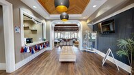 Located at 1000 E 22nd St in Tucson, Arizona, this single-story community clubhouse offers a thoughtfully designed social hub spanning a generous footprint. The entrance area sets an inviting tone with a coffered wood-paneled ceiling, dramatic pendant light fixtures with gold interiors, rich hardwood-style flooring, and a built-in reception window. A tufted bench and merchandise display rack add a polished, welcoming touch. Through a signature white arched passageway, the main lounge and game room unfold beneath soaring wood-beam ceilings lined with warm natural wood planks. This expansive area is anchored by a large sectional sofa, area rug, and wall-mounted TV, while a ping-pong table, pool table, and additional gaming surfaces make it ideal for recreation and socializing. White shiplap-style wainscoting contrasts beautifully with the dark wood flooring throughout. French doors along one wall flood the space with natural light and provide views of and access to an outdoor area. A rustic stone-clad refreshment bar features granite countertops, bar stools, a coffee machine, and open shelving, serving as a casual gathering spot. The overall ambiance blends modern farmhouse aesthetics with comfortable, community-focused functionality.