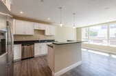 This single-floor unit offers a thoughtfully designed layout with contemporary finishes throughout. The kitchen serves as the centerpiece of the space, showcasing crisp white shaker-style cabinetry, deep granite countertops with a rich, dark veining pattern, and a full suite of stainless steel GE appliances — including a side-by-side refrigerator with ice maker, an electric range, over-the-range microwave, and dishwasher. A generous island with matching granite provides additional prep and seating space, while stylish pendant lighting adds a polished touch overhead. Recessed lighting and wide-plank wood-look flooring run throughout, lending a clean, cohesive aesthetic. The open layout connects the kitchen to an adjoining living or dining area, which benefits from a large multi-pane window that invites abundant natural light and offers views of the surrounding outdoor environment. The bathroom features a walk-in shower with a white surround enclosure and vanity with granite countertop. A dedicated laundry room houses a front-loading washer with connections in place, offering practical in-unit convenience. The overall design is neutral and modern, with warm beige tones, white trim, and quality finishes that create a clean, move-in-ready atmosphere.