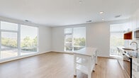 Located at 1200 N Henry St in Alexandria, Virginia, this well-appointed 2-bedroom, 2-bathroom residence offers a thoughtfully designed floor plan with a clean, contemporary aesthetic throughout. The combined kitchen and living area serves as the heart of the home, featuring light wood-tone flooring, recessed lighting, and large windows fitted with horizontal blinds that invite generous natural light and tree-lined outdoor views. The kitchen is outfitted with white cabinetry, stainless steel appliances including a refrigerator, range, and microwave, complemented by warm wood open shelving, subway tile backsplash, and light quartz countertops. A hallway with recessed lighting connects the main living area to the private quarters, where two bedrooms are finished with plush neutral carpeting and crisp white walls. The primary bedroom is a well-sized retreat with access to a dedicated closet. Both bathrooms continue the modern palette, with the main bath showcasing a sleek white vanity, quartz countertop, large mirror, and contemporary fixtures. Three closets throughout the unit provide ample storage. The consistent use of white tones, light flooring, and modern hardware across all spaces creates a bright, cohesive ambiance ideal for comfortable urban living.